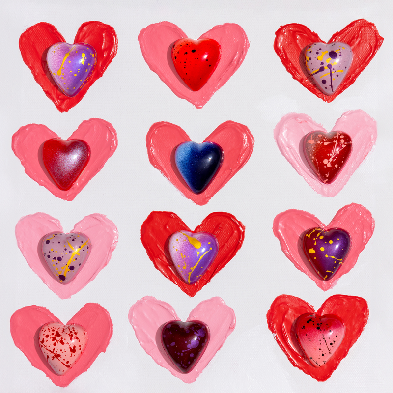 12 multi-colored heart-shaped chocolates displayed in a 3x4 grid. The chocolates are a variety of colors(pink, purple, blue) and placed on pink or red painted hearts to highlight the color and design.